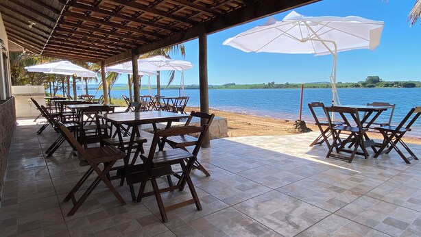 An outdoor patio with wooden tables and chairs, white umbrellas, and a view of a lake. - Country house, heated pool, beach, tennis court, barbecue, playground, fishing, pets allowed. (Barão de Antonina)