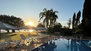 A swimming pool with lounge chairs and umbrellas, surrounded by palm trees and a sunset in the background. - Country house, heated pool, beach, tennis court, barbecue, playground, fishing, pets allowed. (Barão de Antonina)