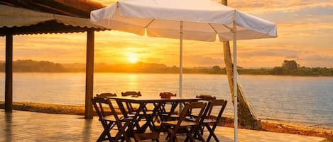 A patio with a table and chairs under a white umbrella, overlooking a body of water with a sunset in the background.