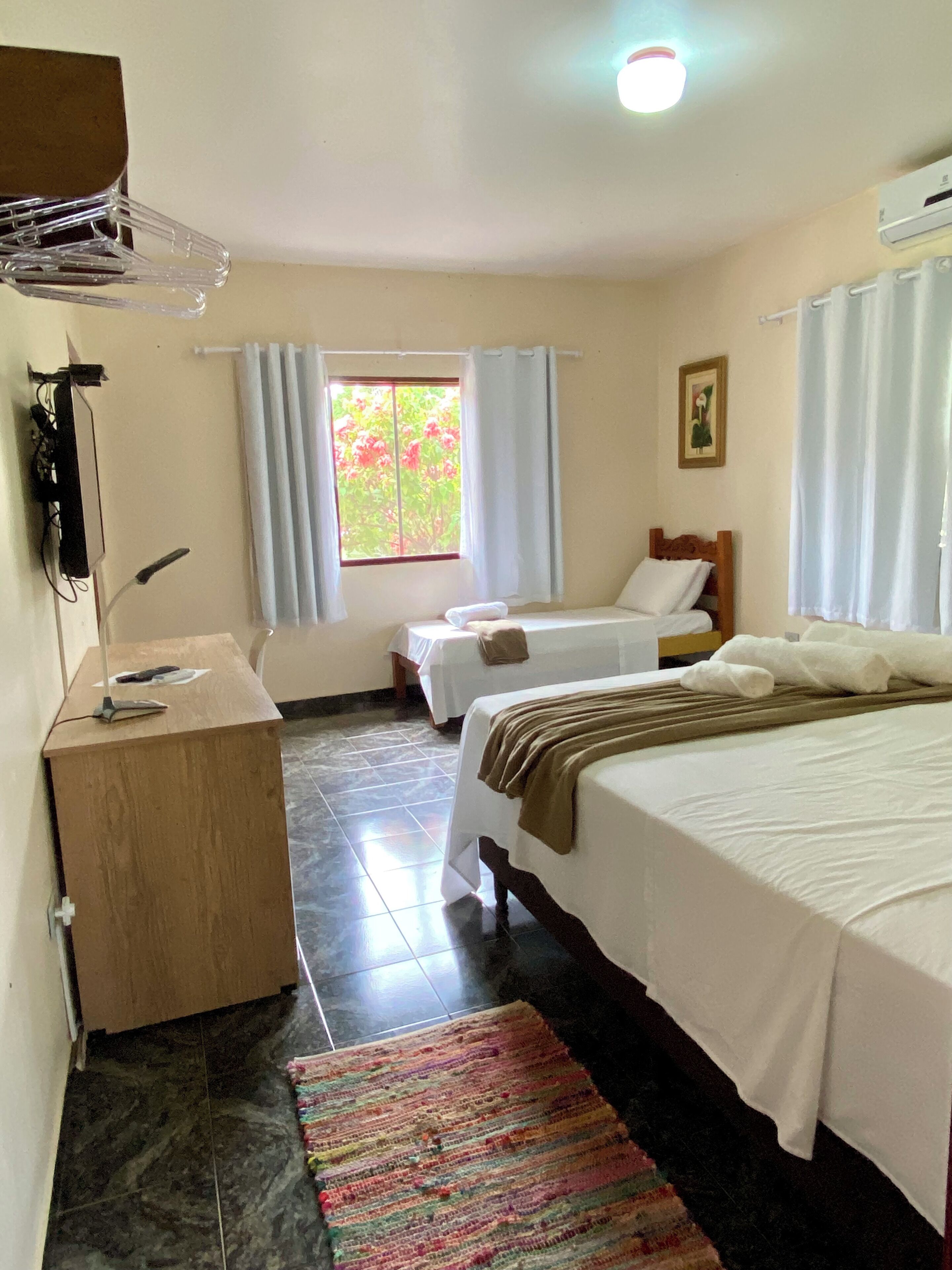 A hotel room with two beds, a desk, a window with curtains, a tiled floor, and a colorful rug.