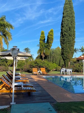 Uma área à beira da piscina com espreguiçadeiras de madeira, um pergolado com uma luminária e uma alta árvore cipreste.