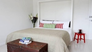 Une chambre à coucher avec un lit, une commode en bois, un vase de fleurs et un tabouret rouge.