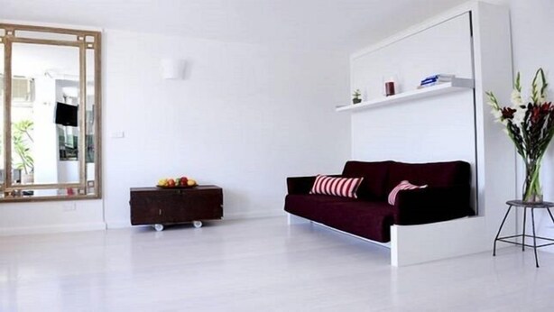 Un salon minimaliste avec un canapé rouge foncé, une petite commode en bois sur laquelle sont disposés des fruits et un miroir.