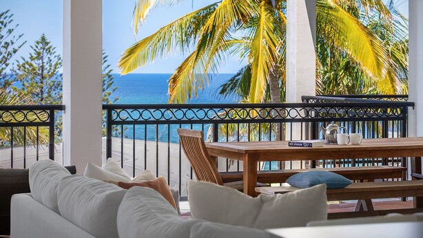A balcony with a view of the ocean, a wooden table with a book and a teapot, and a sofa with pillows.