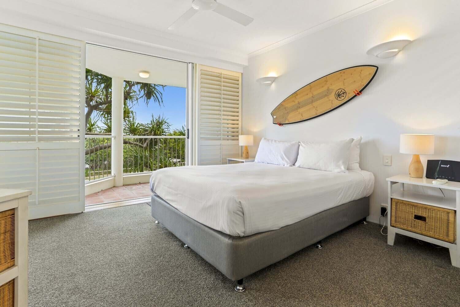 Een slaapkamer met een groot bed, een surfboard aan de wand, een balkon met uitzicht op palmbomen en een plafondventilator.