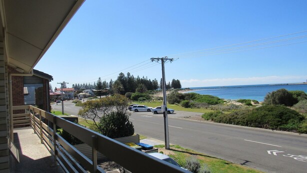 A coastal road with parked cars, a pedestrian path, and a view of the sea and a pier.