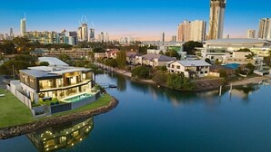 A modern residential area with a large house featuring a swimming pool, surrounded by a calm lake, with a city skyline in the background.