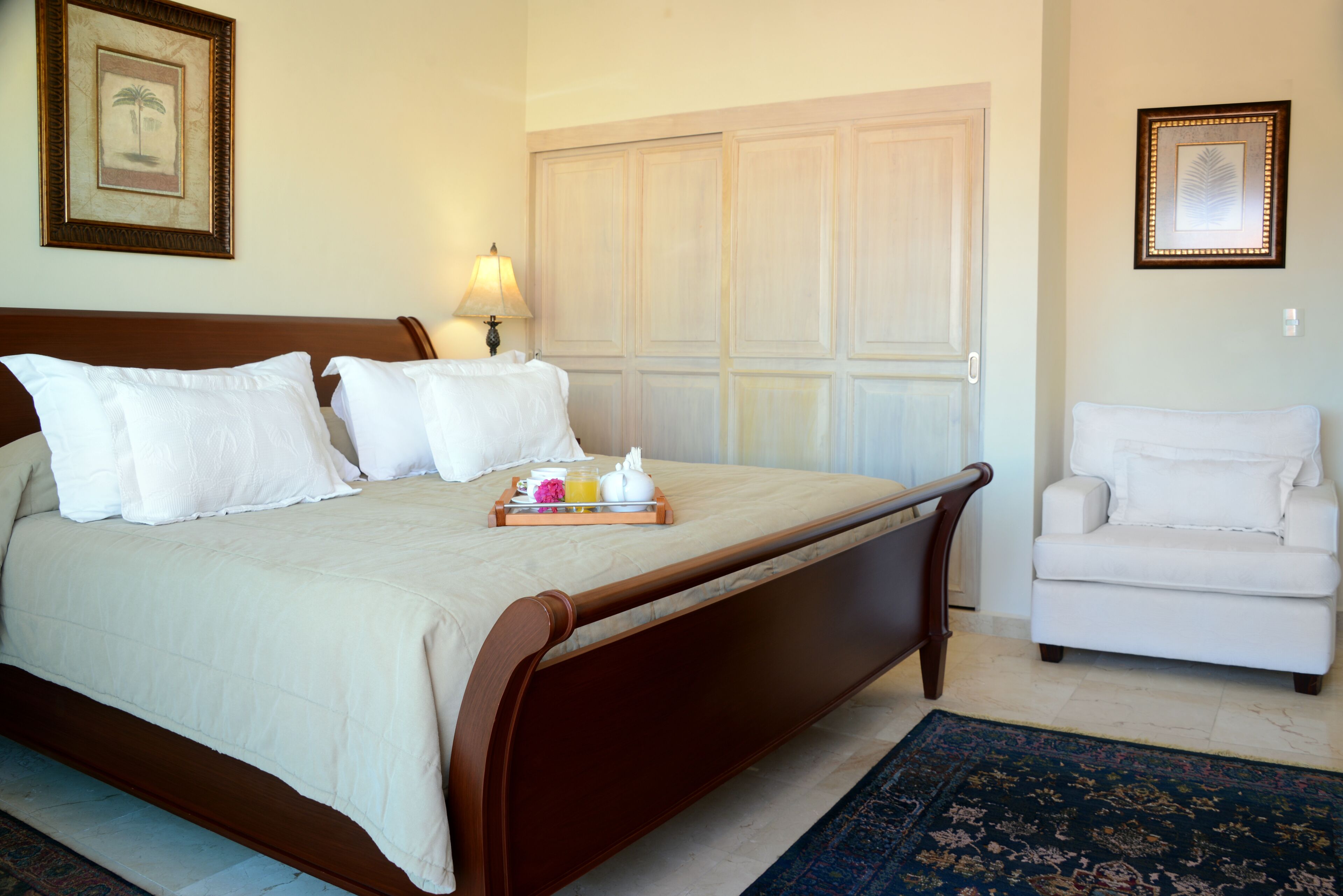 A neatly made bed with white linens, a bedside table with a tray of breakfast items, and a white armchair in a hotel room.