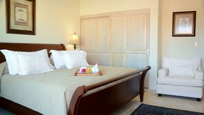 Un lit bien fait, avec du linge de lit blanc, une table de chevet sur laquelle se trouve un plateau avec des articles pour le petit-déjeuner, et un fauteuil blanc dans une chambre d’hôtel.