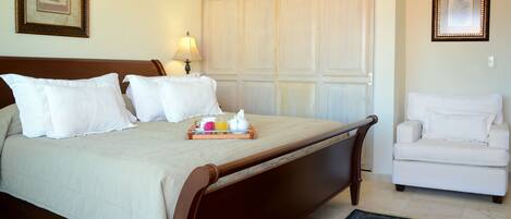 A neatly made bed with white linens, a bedside table with a tray of breakfast items, and a white armchair in a hotel room.