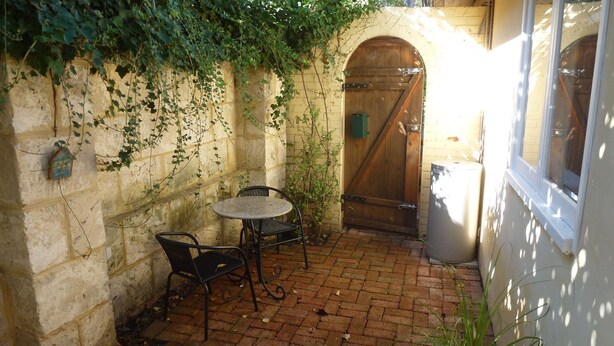 Un patio con suelo de ladrillo, una mesa redonda con dos sillas, una puerta de madera y una caja verde pequeña en la pared.