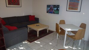 Una sala de estar con un sofá gris, cojines rojos, una mesa de centro de madera y un televisor en la pared. Hay una zona de comedor con una mesa blanca y sillas marrones.