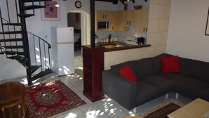 Una sala de estar con un sofá gris, cojines rojos, una mesa de centro de madera, un tapete rojo estampado, una escalera y una zona de cocina con armarios de madera y un microondas.