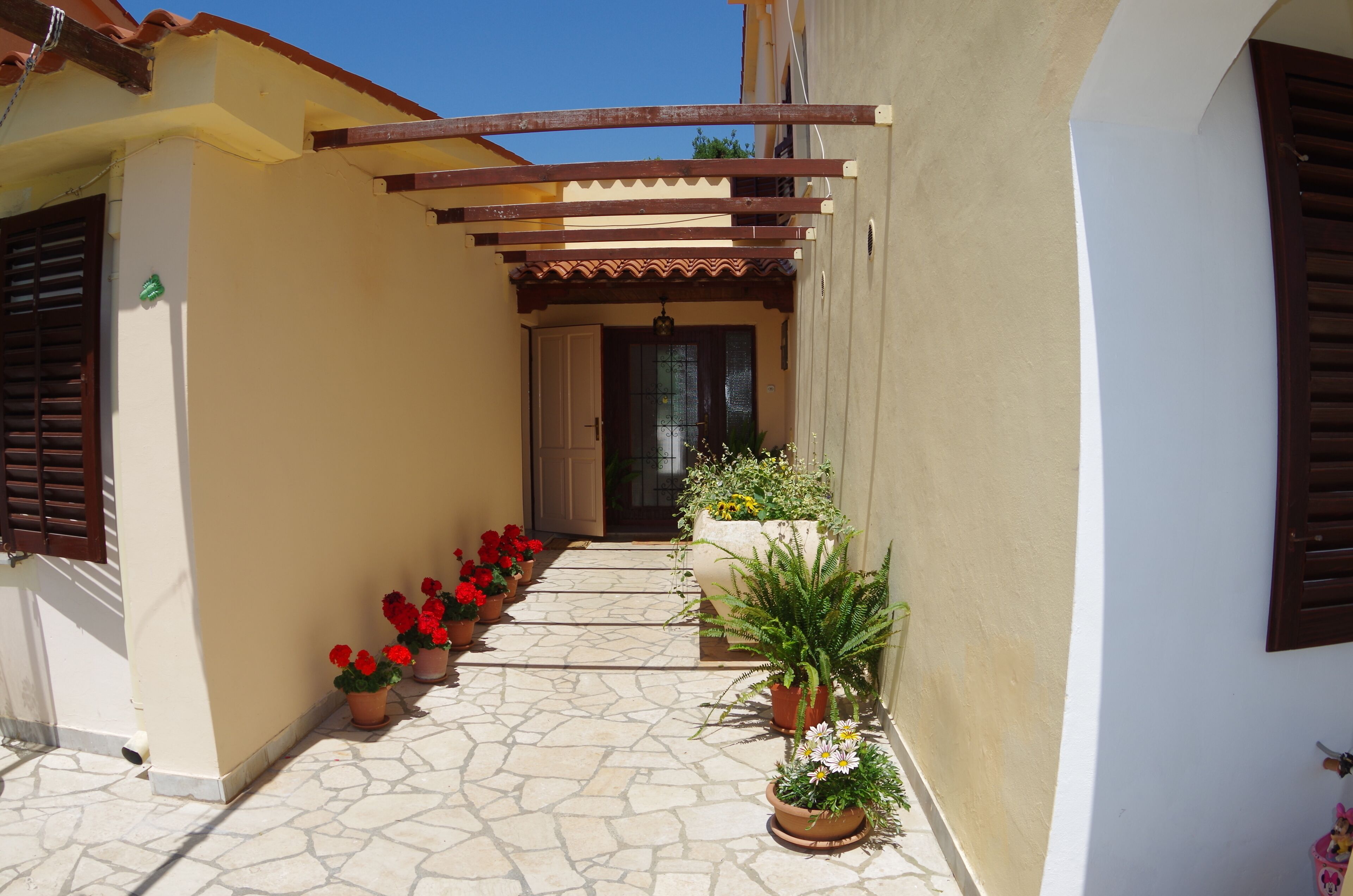 A narrow courtyard with a tiled floor, potted plants, and a wooden pergola.