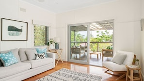 A spacious living room with a white sofa, a patterned rug, and a large sliding glass door leading to an outdoor patio with wooden furniture. - The White Rabbit - with studio & covered deck (Byron Bay)