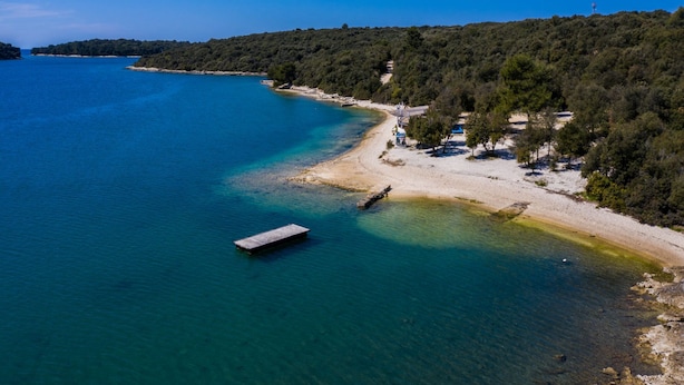 An aerial view of a coastal area with a dock, clear blue waters, and a sandy beach with trees.