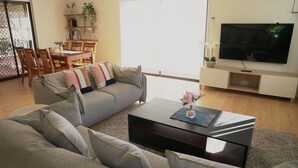 A modern living room with a grey sofa, a coffee table, and a flat-screen TV. There is a dining area with a table and chairs, and a bookshelf with plants.