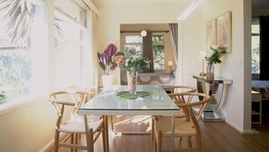 A dining area with a glass table, wooden chairs, and a view of a bedroom with a bed and curtains.