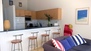 Una cocina moderna con armarios de madera, encimera blanca y taburetes de bar. En la sala de estar, hay un sofá gris con almohadones a rayas y una manta blanca y roja a rayas.