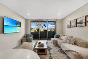 Ett modernt vardagsrum med en stor TV, bekväma sittplatser, ett soffbord och utsikt över en tropisk strand genom glasdörrarna.