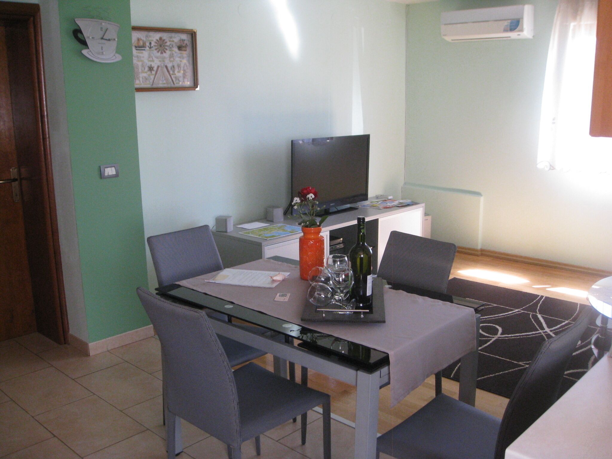 A dining area with a glass table, four chairs, a television, and a small cabinet.