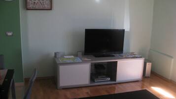 A living room with a flat-screen TV on a white cabinet, a small table with a globe and papers, and a black rug with geometric patterns.
