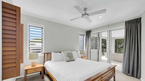 Un dormitorio con cama, ventilador de techo, mesita de noche, ventana con persianas y vistas al exterior.