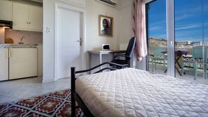 Ein Schlafzimmer mit Bett, Schreibtisch, Stuhl, Fenster mit Aussicht und einer Küchenzeile.