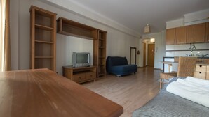 Ein Zimmer mit Holztisch, einem Bett mit grauer Decke, einem Fernseher auf einem Holzständer, einem blauen Sofa und einer Küchenzeile mit Holzausstattung.