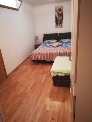A bedroom with a wooden floor, a bed with a striped bedspread, a nightstand with a lamp, and a painting on the wall.