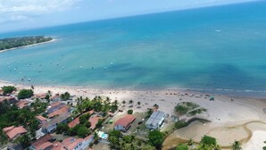 Vista aérea de uma praia com águas turquesa cristalinas, uma faixa de areia e uma pequena área residencial com edifícios de telhado vermelho.