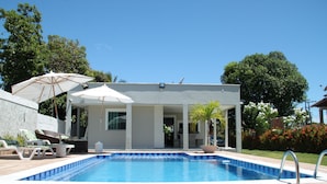 Piscina com espreguiçadeiras e guarda-sóis, cercada por um jardim e árvores.