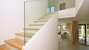 Un intérieur moderne avec un escalier à garde-corps en verre, un espace de vie comprenant un canapé et des rideaux, et une vue sur l’extérieur.
