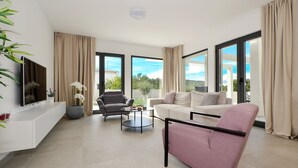 Un salon moderne avec un canapé, un fauteuil et une table basse. On y trouve un téléviseur à écran plat posé sur une console blanche, ainsi qu’une grande fenêtre avec des rideaux.
