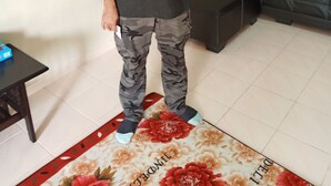 Una persona de pie en una habitación con un sofá oscuro, una mesita pequeña y un tapete estampado con motivos florales.