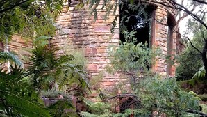 Un bâtiment en pierre, d’aspect rustique et empreint d’histoire, entouré d’une végétation luxuriante et de fougères.