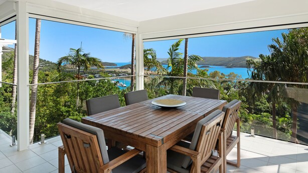 Une table en bois, entourée de chaises, est placée sur un balcon qui offre une vue panoramique sur une région côtière, avec des palmiers et une étendue d’eau.