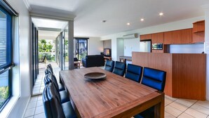 Une cuisine moderne avec des meubles en bois, un coin repas avec une table en bois et des chaises bleues, et une grande porte-fenêtre coulissante donnant sur un espace extérieur.