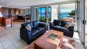 Un salon moderne avec deux canapés en cuir noir, une table basse en bois et un coin repas comprenant une table et des chaises en bois. De grandes portes-fenêtres coulissantes donnent sur un balcon offrant une vue sur la mer.