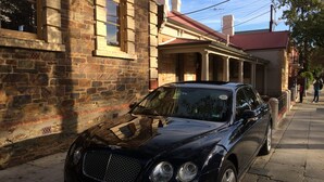 A black car parked on a sidewalk next to a stone building with arched windows and a red roof. - Friendly meeting chapel (North Adelaide)