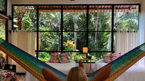 A room with a large window, a hammock, two beds, and a small table with a lamp.