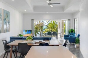 A modern room with a dining area, a sofa, and a TV. There is a view of palm trees through large windows.