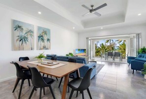 A modern living room with a dining area, a ceiling fan, and a large window with a view of palm trees.