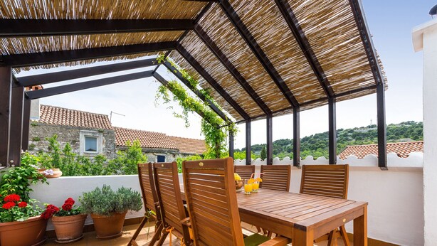 Un porche cubierto con una mesa y sillas de madera, plantas en macetas y vistas de edificios al fondo.