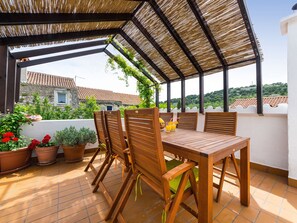 Une terrasse couverte avec une table et des chaises en bois, des plantes en pot et une vue sur les bâtiments en arrière-plan.