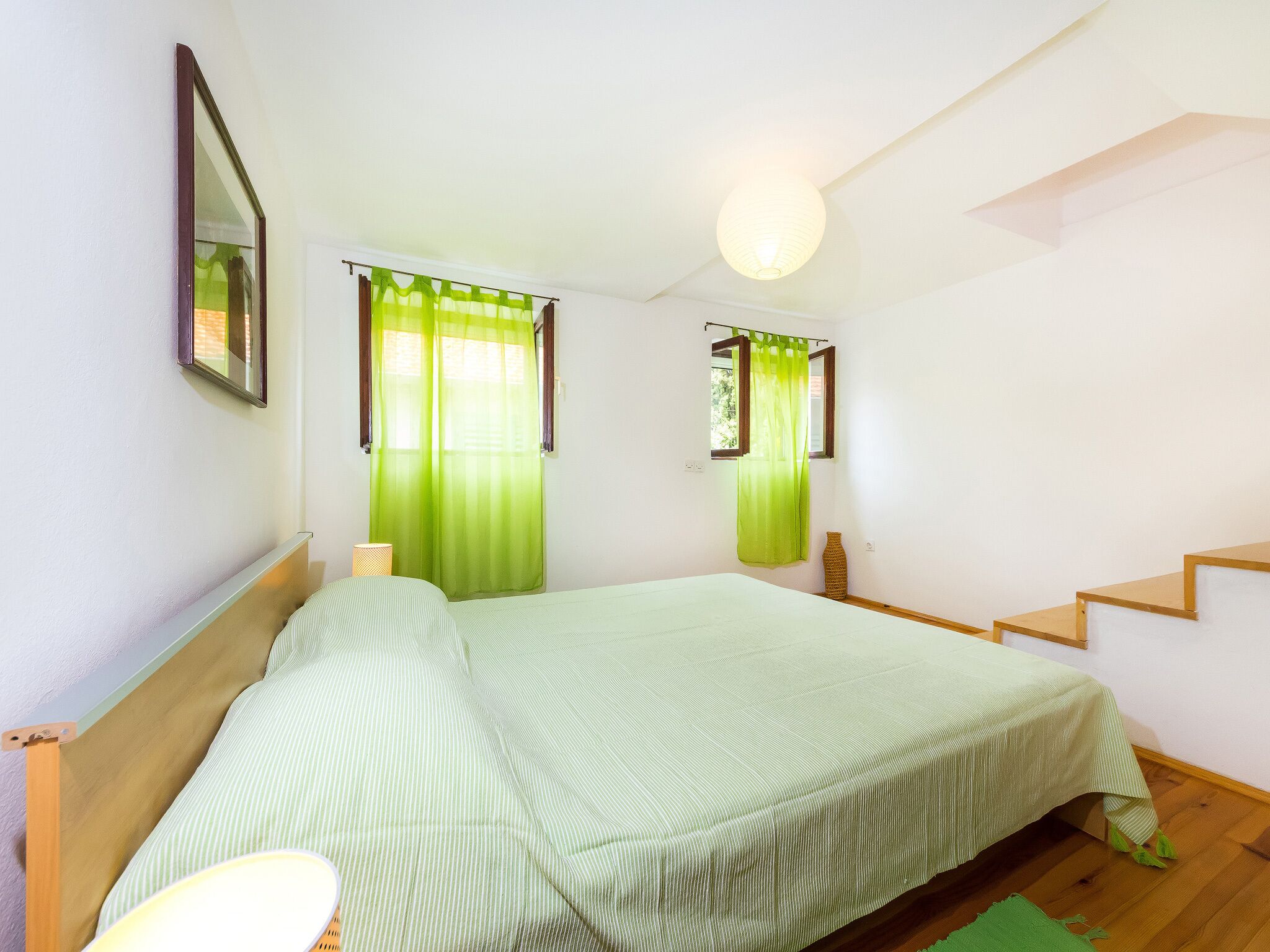 Ein Schlafzimmer mit Bett, Nachttisch, Lampe, einem Fenster mit Vorhängen, einem Spiegel und einer Treppe.