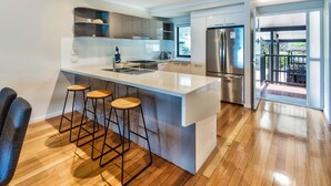 A modern kitchen with a central island, bar stools, stainless steel appliances, and a view to an outdoor balcony. - Shorelines 26, Seaview Apartment on Hamilton Island (Hamilton Island)