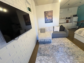 Ein modernes Wohnzimmer mit Flachbildfernseher, grauem Sofa, einem gemusterten Teppich und einer Küche im Hintergrund.