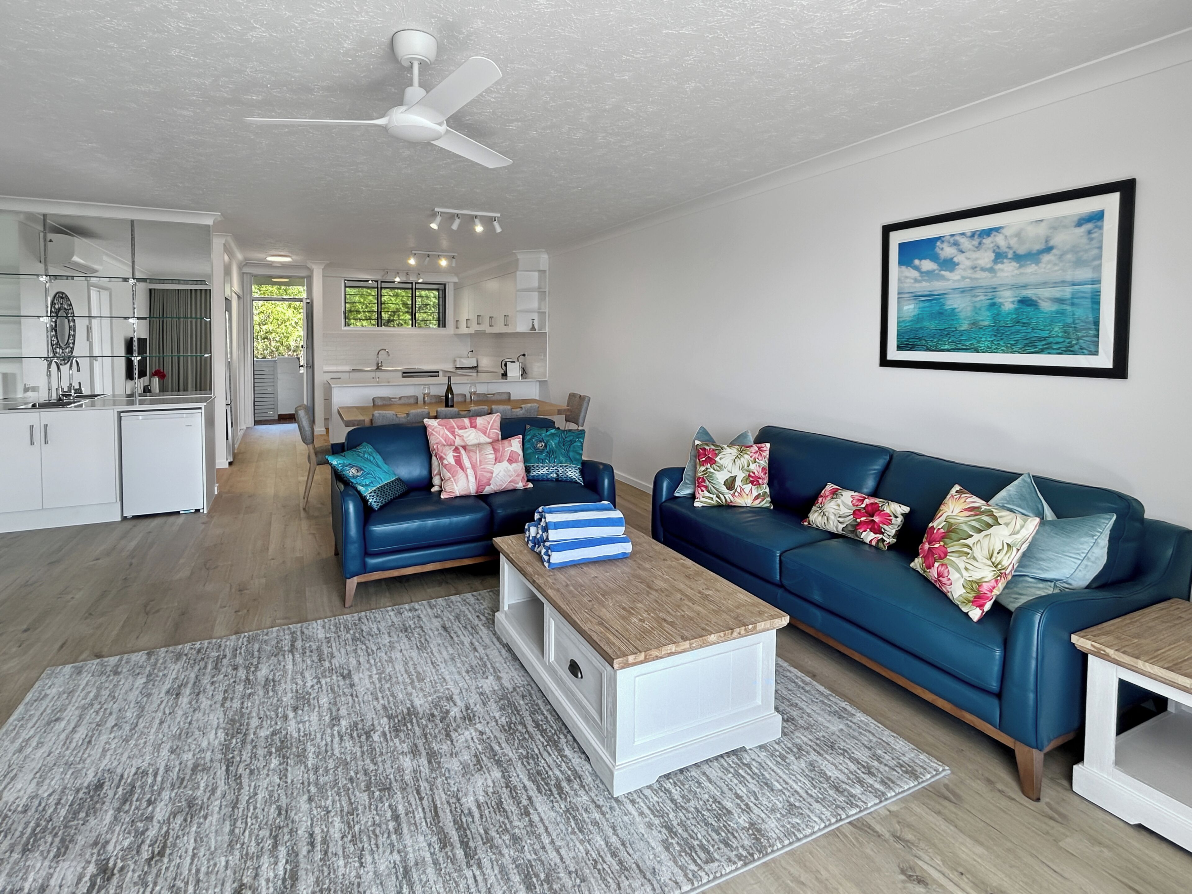 A modern living room with a blue sofa, a coffee table, and a rug. There is a kitchen in the background with white cabinets and a dining area.