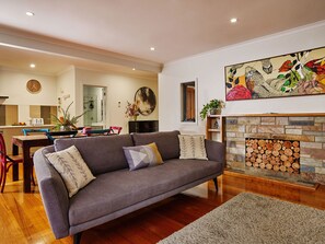 A living room with a grey sofa, wooden flooring, a fireplace with logs, and a dining area with chairs and a table.